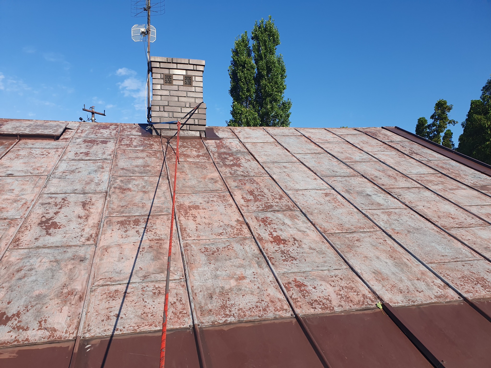 Stary dach pokryty metalową dachówką z widocznymi śladami korozji i zabrudzeń, na tle błękitnego nieba z kominem i anteną.