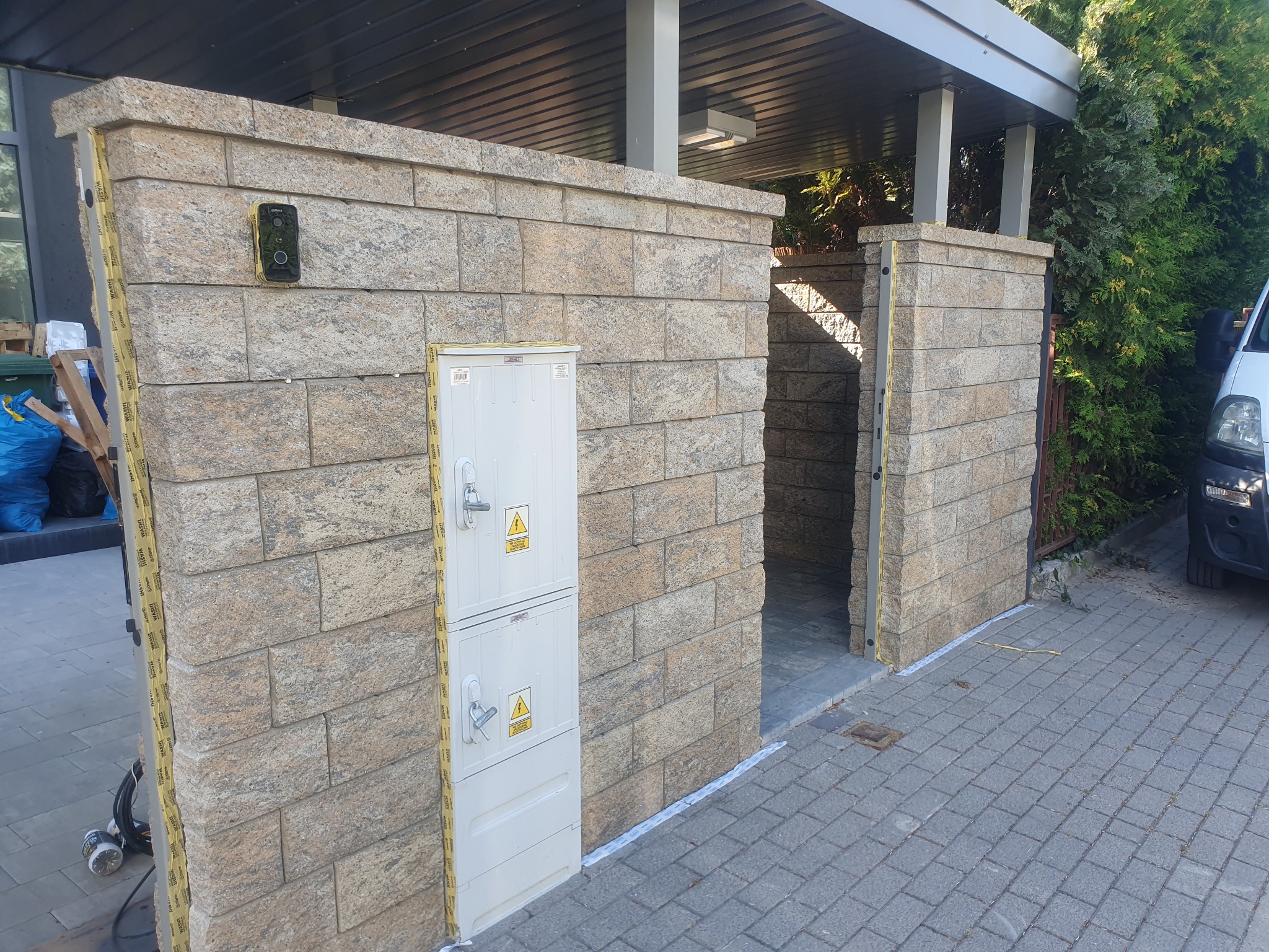 Zewnętrzna konstrukcja z bloków kamiennych z zainstalowaną skrzynką elektryczną i wideodomofonem, widoczne ślady prac wykończeniowych, w tle fragment zaparkowanego samochodu.