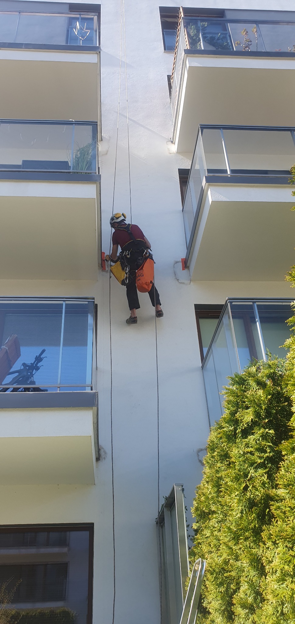 Pracownik na linach, w kasku i z narzędziami, czyści elewację budynku wielopiętrowego, widoczne balkony z szklanymi balustradami i zieleń.