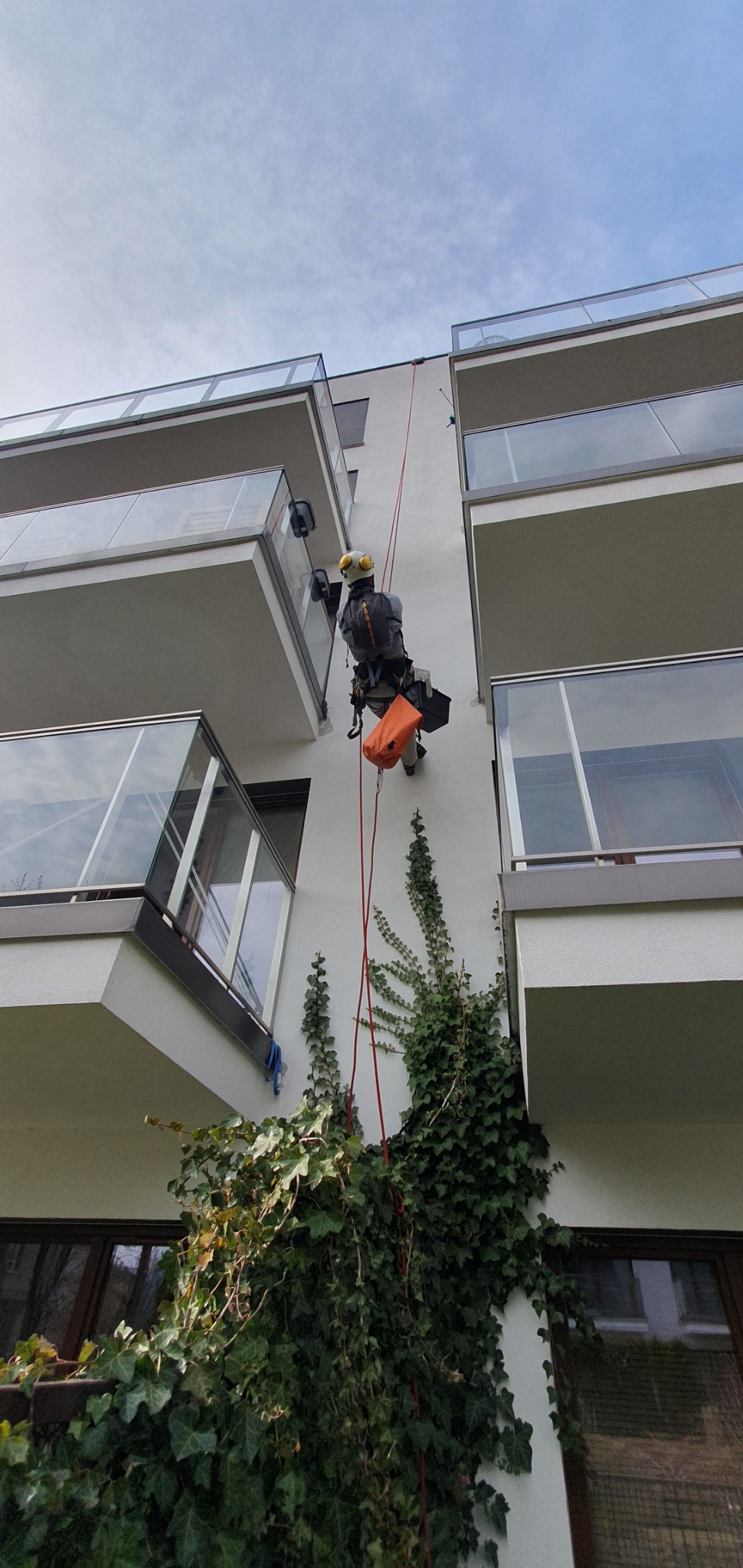 Pracownik na linie zabezpieczonej, w kasku i z torbą narzędziową, wykonuje prace na elewacji budynku z balkonami, w tle widoczna ściana porośnięta bluszczem.