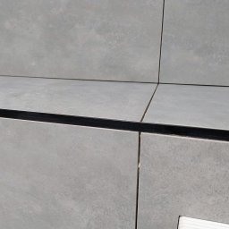 Usługi glazurniczy Kraków Piotr Kula - Szare, matowe płytki ceramiczne ułożone na ścianie i blacie, z widoczną czarną listwą wykończeniową. Minimalistyczny design, precyzyjne fugi.