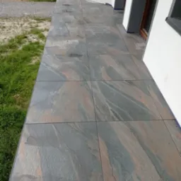 Wykończony taras z szarych, wielkoformatowych płytek imitujących kamień, widok z boku, z fragmentem trawnika i elewacji budynku.