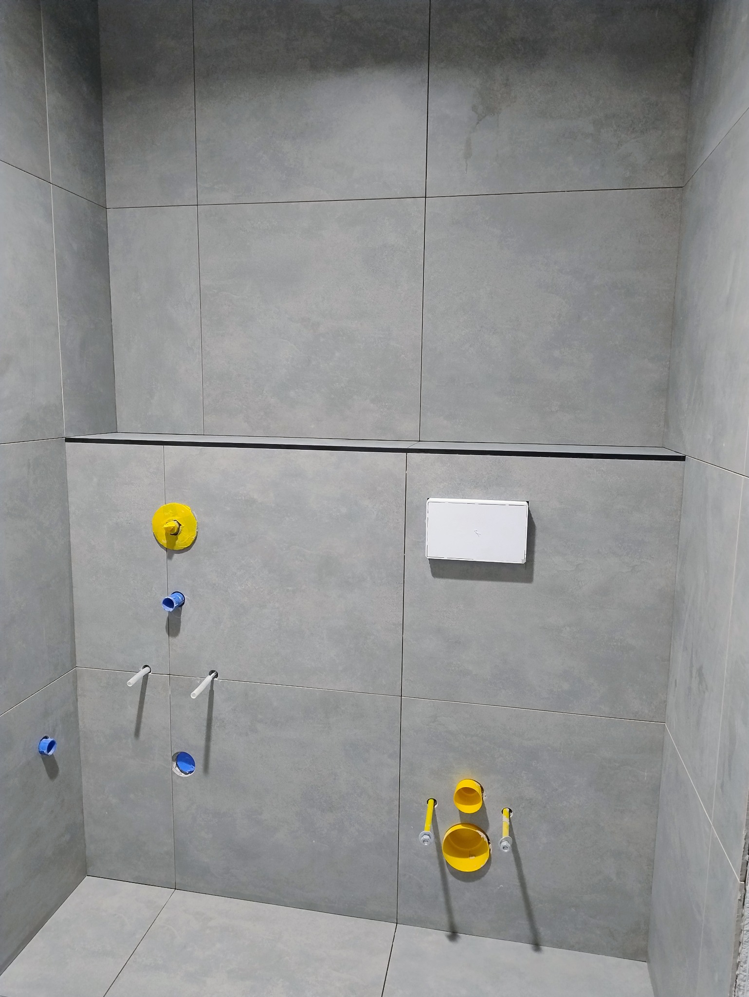 Szara łazienka w trakcie wykańczania: widoczne płytki, instalacja wodna z żółtymi i niebieskimi elementami, oraz biały przycisk spłuczki. Minimalistyczny design.
