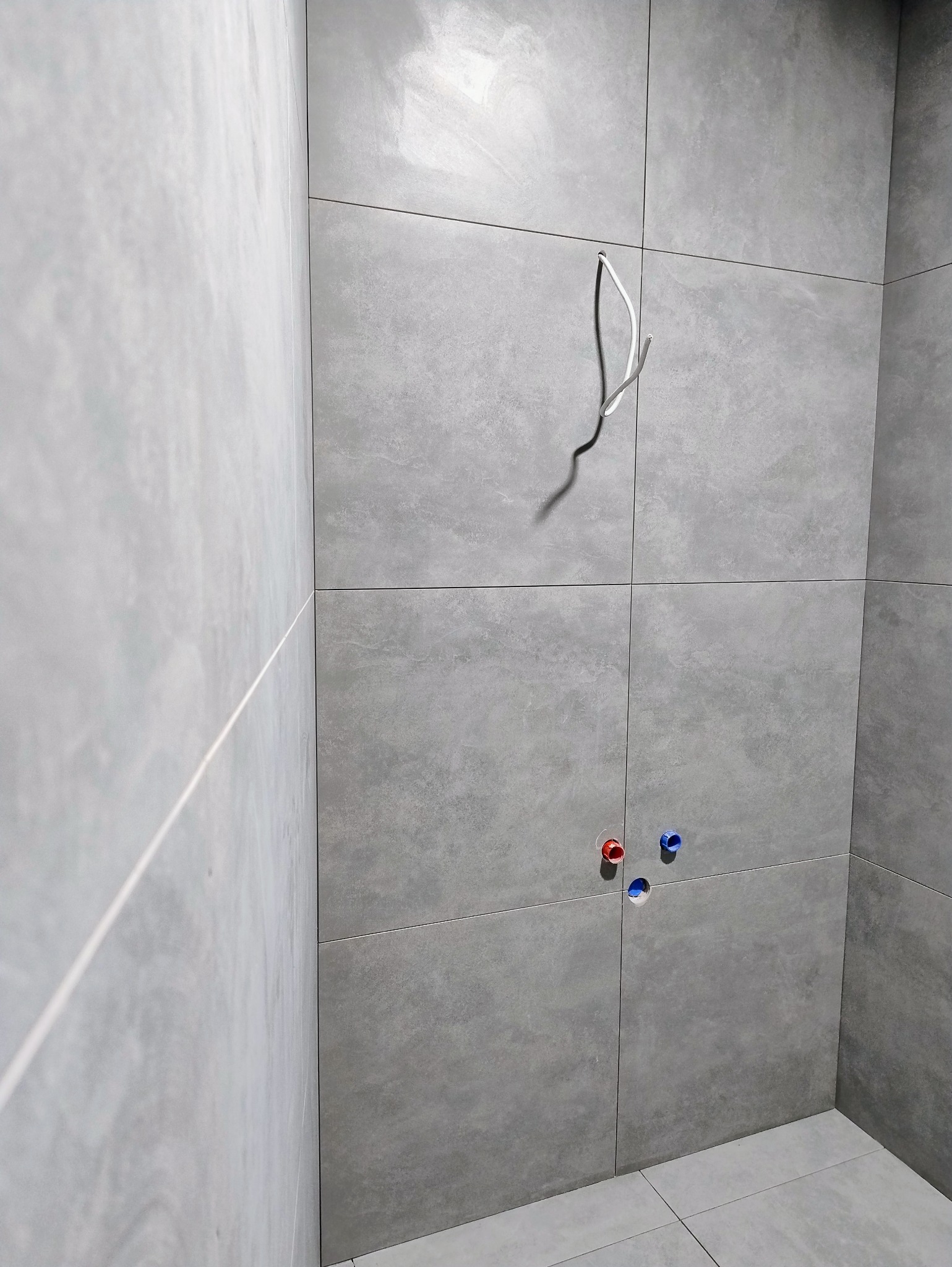 Ściana w łazience wyłożona szarymi płytkami, z widocznymi wystającymi przewodami elektrycznymi i rurami z ciepłą i zimną wodą. Minimalistyczny design.