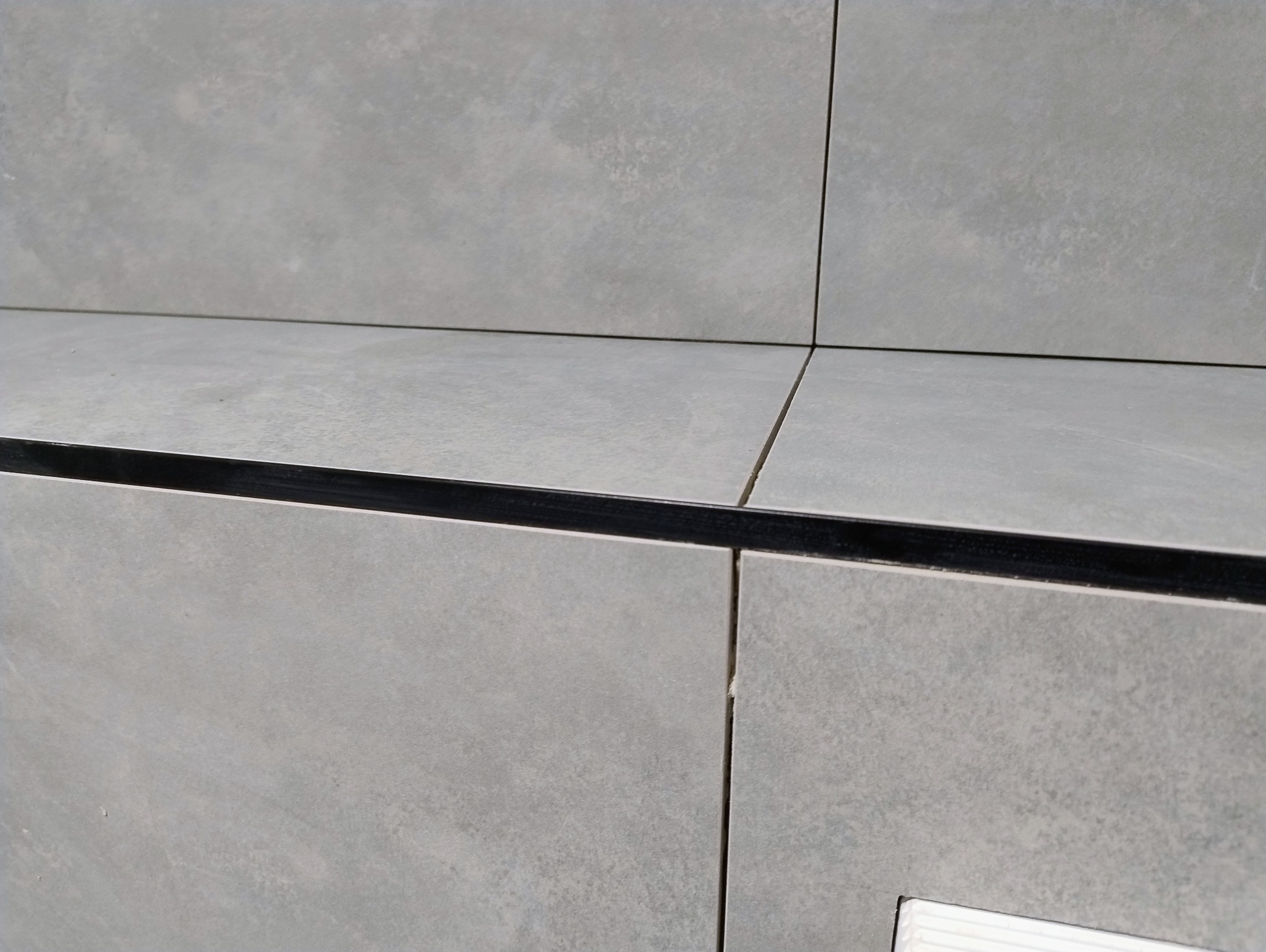 Szare, matowe płytki ceramiczne ułożone na ścianie i blacie, z widoczną czarną listwą wykończeniową. Minimalistyczny design, precyzyjne fugi.