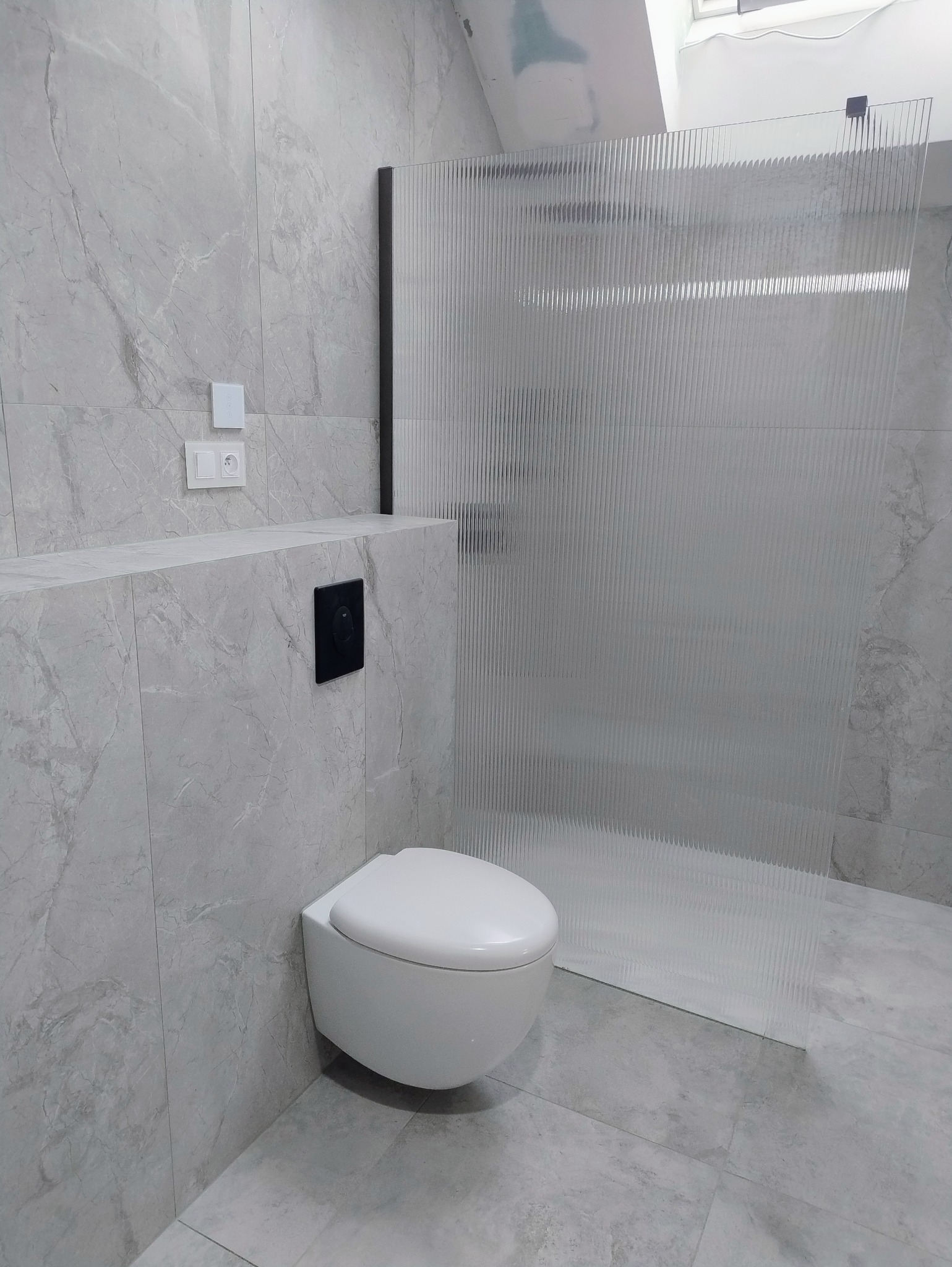 Nowoczesna łazienka w szarościach: podwieszana toaleta, szklana kabina prysznicowa z prążkowaną szybą, płytki imitujące marmur na ścianach i podłodze.