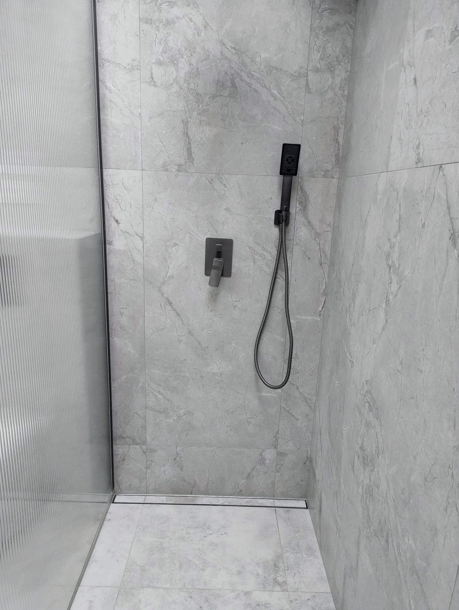 Nowoczesna kabina prysznicowa z szarymi płytkami imitującymi marmur, czarną armaturą i odpływem liniowym. Minimalistyczny design, eleganckie wykończenie.