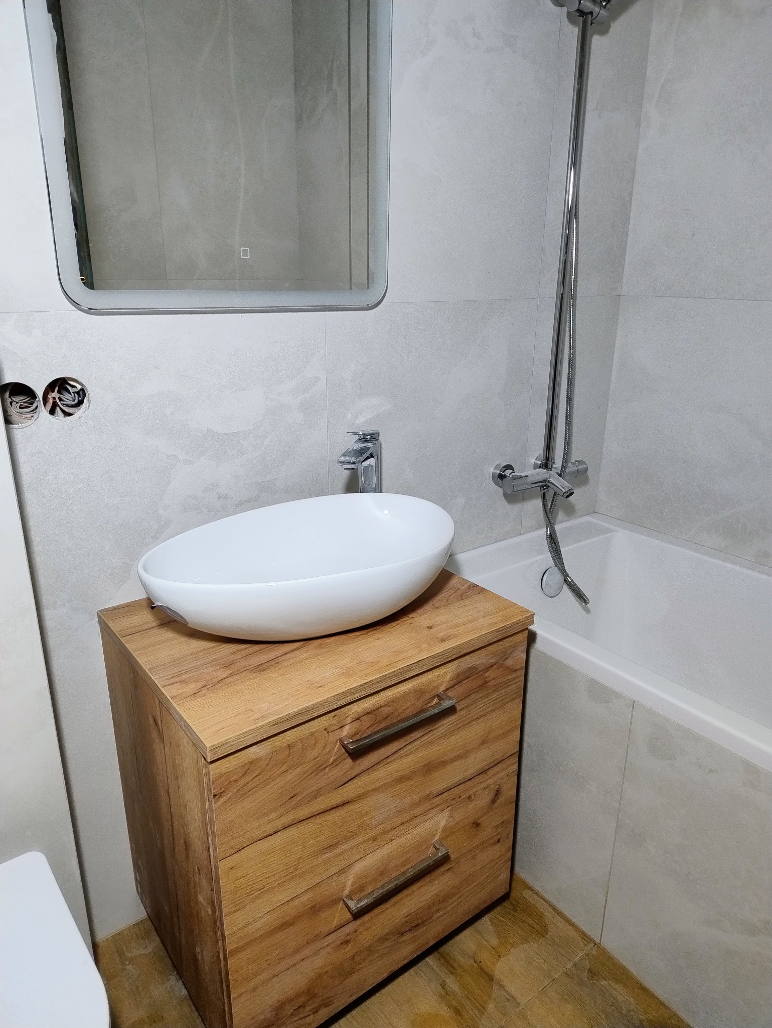 Nowoczesna łazienka w Krakowie: owalna umywalka na drewnianej szafce, lustro z podświetleniem i szara glazura. Minimalistyczny design i funkcjonalność.