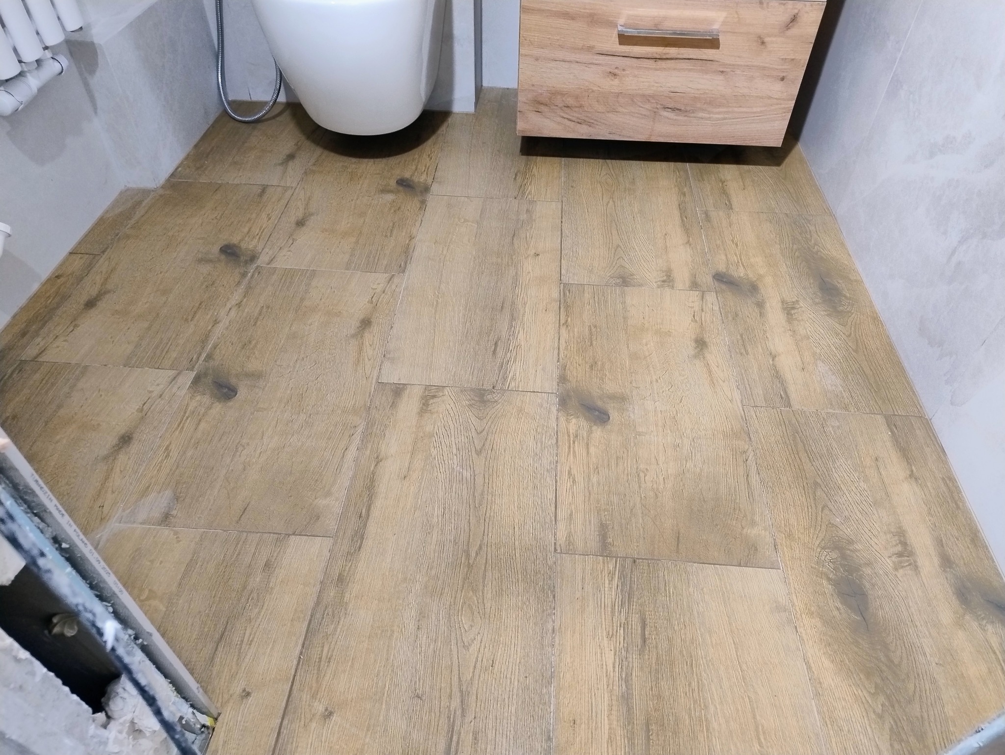 Ułożona podłoga z płytek imitujących drewno w łazience, widoczna toaleta i szafka pod umywalką. Jasne płytki na ścianach.