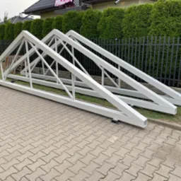 Dwie białe, stalowe konstrukcje kratowe leżą na kostce brukowej, opierając się o trawnik i czarny płot. W tle zielone tuje i fragment dachu domu.