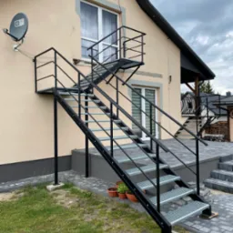 Zewnętrzne schody stalowe z platformą, prowadzące do drzwi na piętrze. Konstrukcja pomalowana na czarno, z kratowymi stopniami, zamontowana przy elewacji domu.