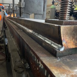 Fix&Tune - Piotr Szumlak - Spawanie długiego elementu stalowego w warsztacie, widoczne iskry i ślady spawania na metalu. W tle butla z gazem i osoba w odzieży roboczej.