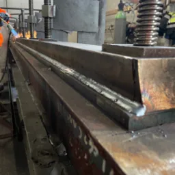 Spawanie długiego elementu stalowego w warsztacie, widoczne iskry i ślady spawania na metalu. W tle butla z gazem i osoba w odzieży roboczej.