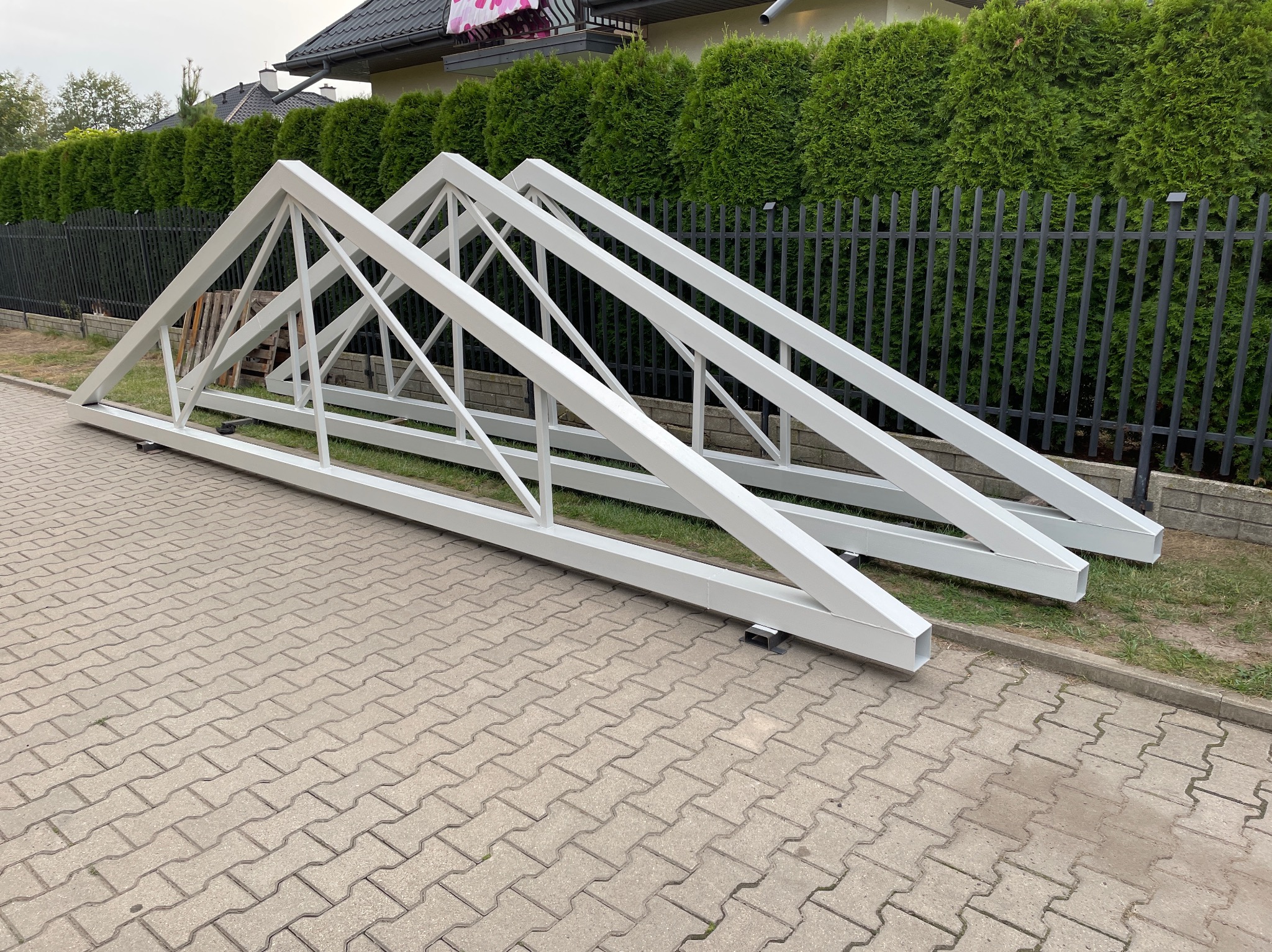 Dwie białe, stalowe konstrukcje kratowe leżą na kostce brukowej, opierając się o trawnik i czarny płot. W tle zielone tuje i fragment dachu domu.