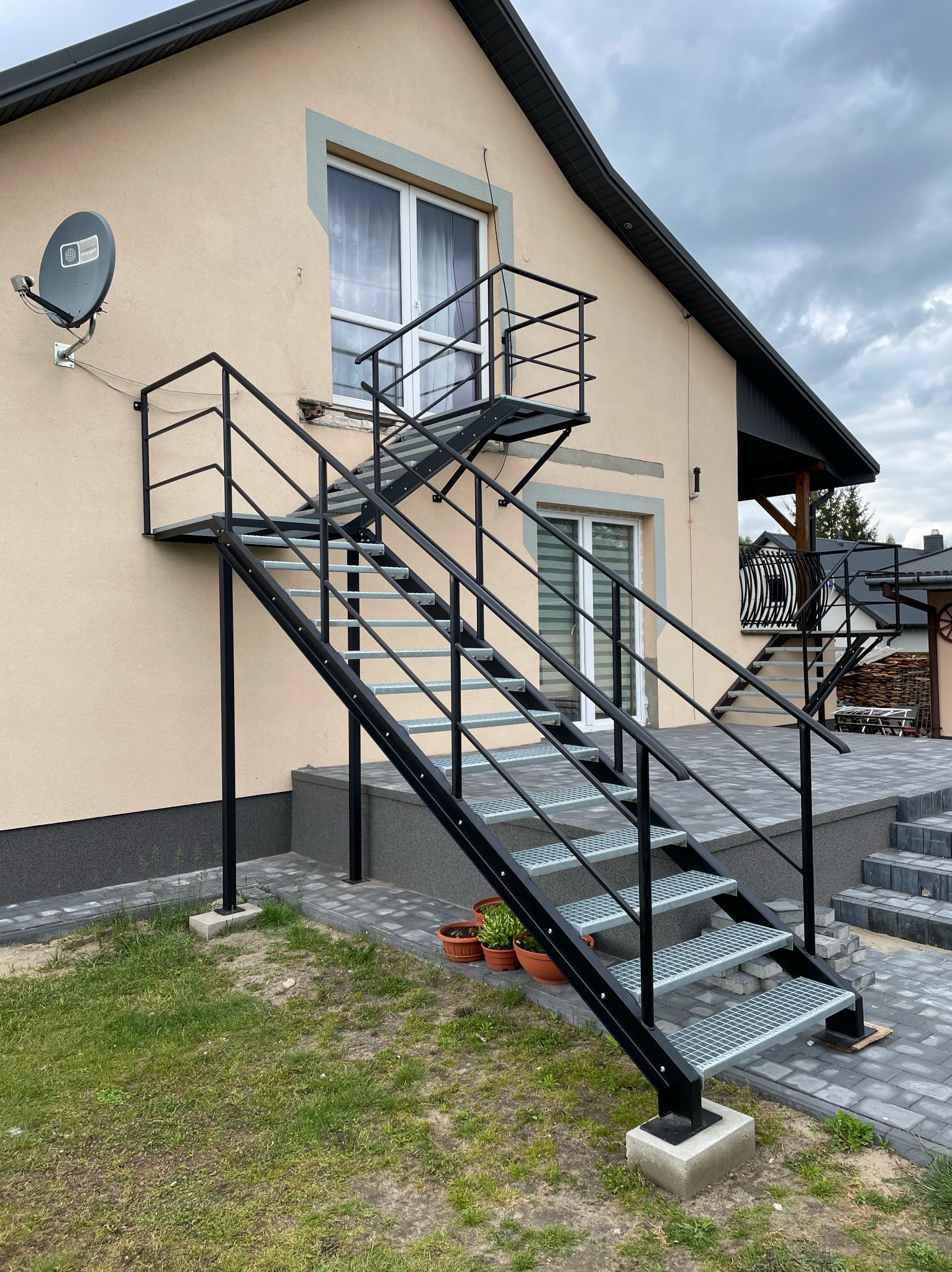 Zewnętrzne schody stalowe z platformą, prowadzące do drzwi na piętrze. Konstrukcja pomalowana na czarno, z kratowymi stopniami, zamontowana przy elewacji domu.