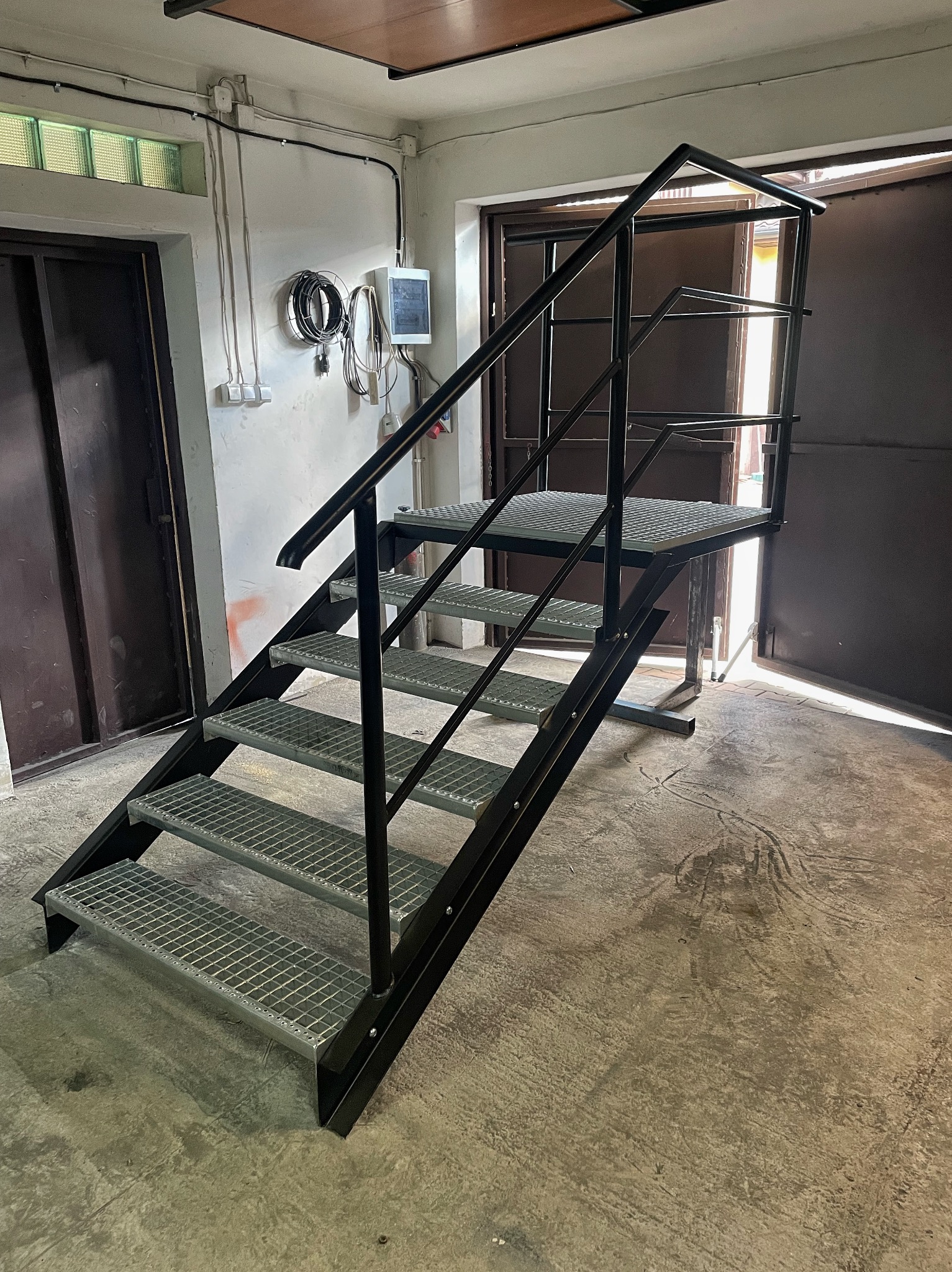 Metalowe schody z podestem i poręczą, z kratowymi stopniami, w hali. Widoczne instalacje elektryczne na ścianie i otwarte drzwi w tle.