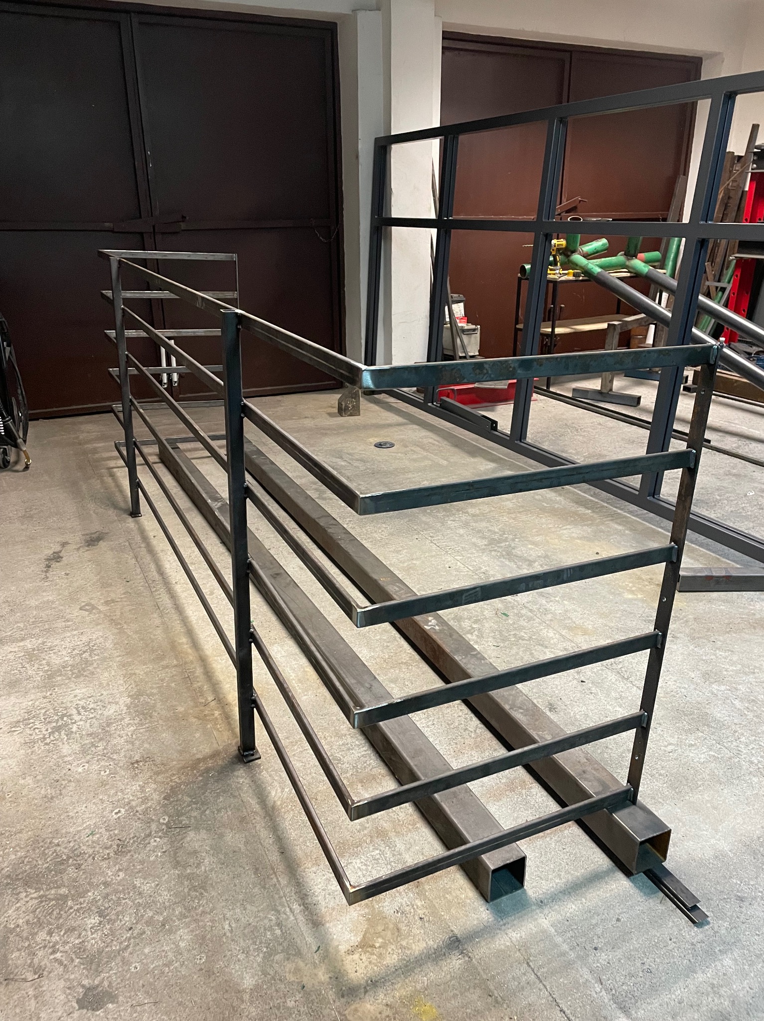 Metalowa konstrukcja schodów z barierką w warsztacie, spawana z profili, surowe wykończenie, widoczne ślady spawania, betonowa posadzka, ciemne drzwi w tle.