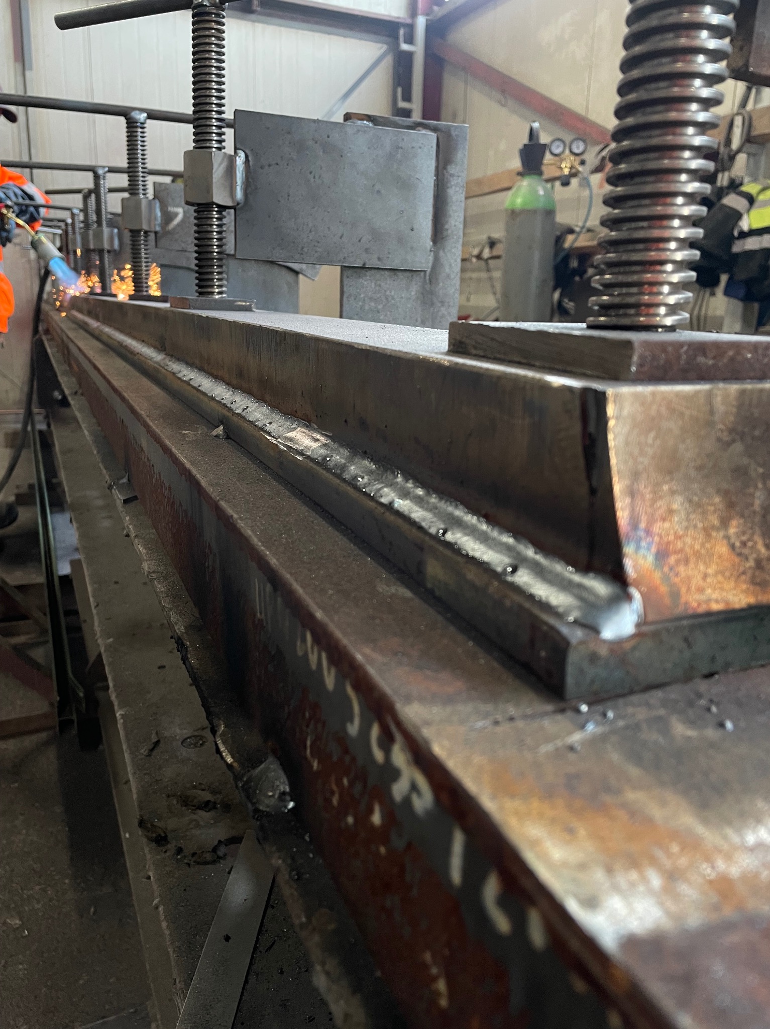 Spawanie długiego elementu stalowego w warsztacie, widoczne iskry i ślady spawania na metalu. W tle butla z gazem i osoba w odzieży roboczej.