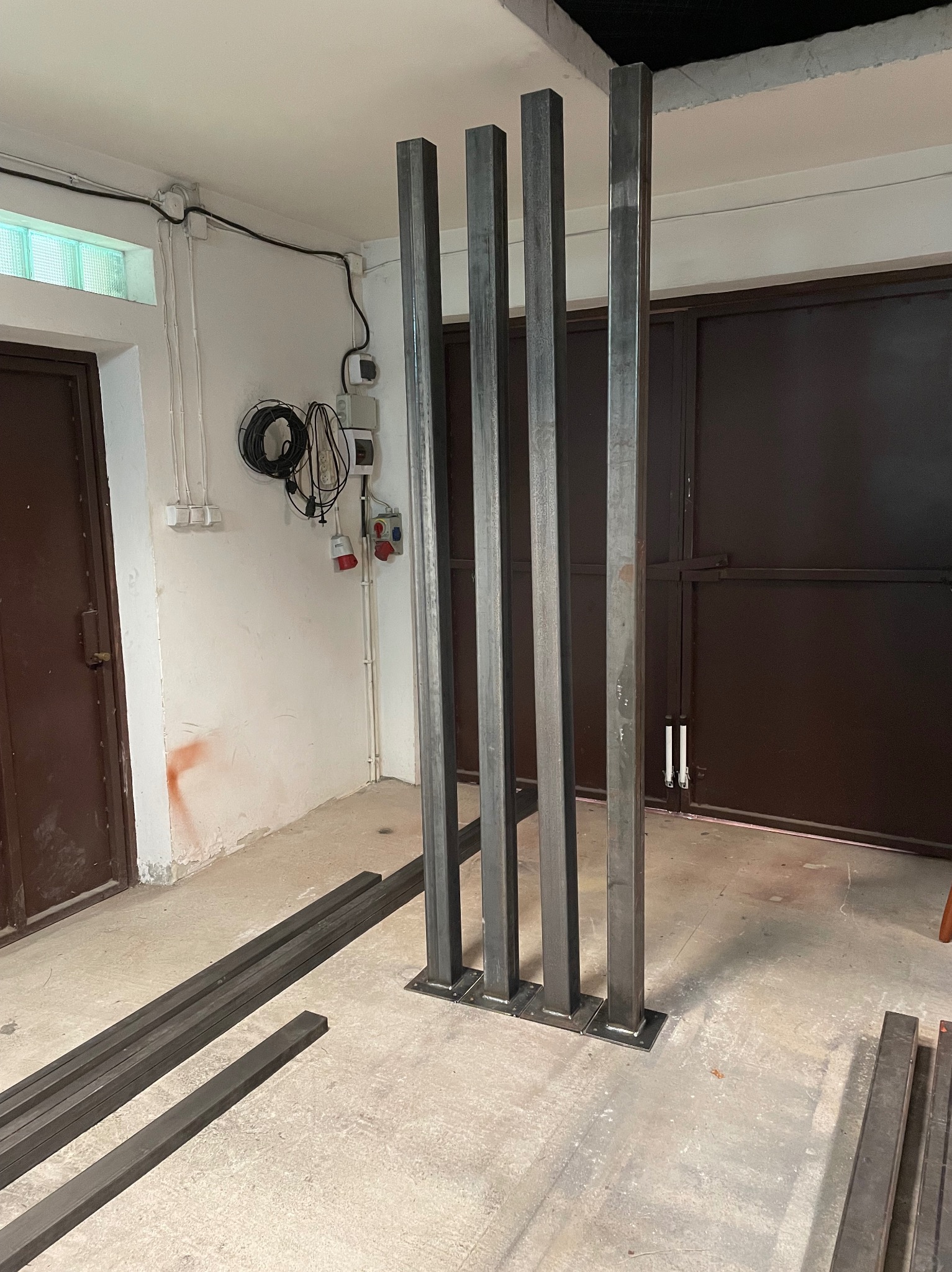 Surowe, stalowe słupy konstrukcyjne w warsztacie, ustawione pionowo, z widocznymi spawami u podstawy. Dodatkowe elementy leżą na podłodze w tle.