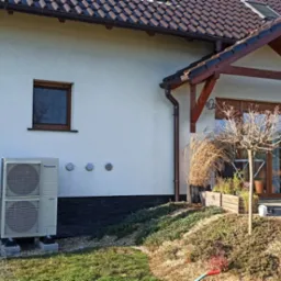 Zewnętrzna jednostka pompy ciepła Panasonic zamontowana przy ścianie domu z widocznymi trzema otworami wentylacyjnymi, z przodu trawnik i element instalacji ogrodowej.