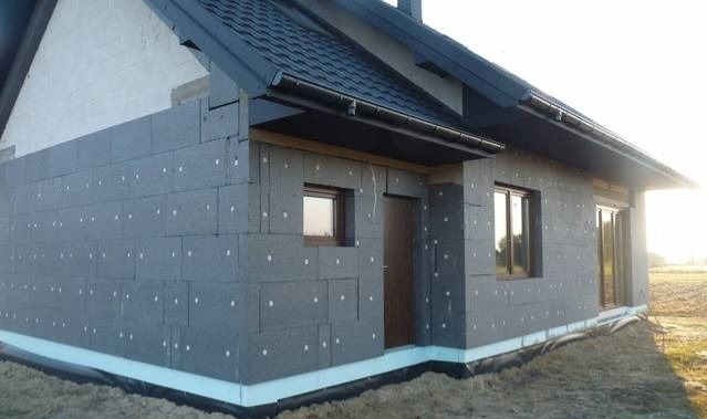 Dom jednorodzinny w trakcie ocieplania elewacji styropianem grafitowym, widoczne kołki mocujące i niezabezpieczona podstawa budynku.