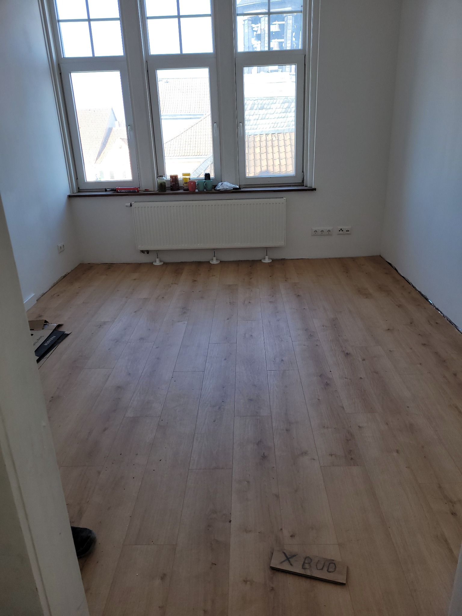 Pusty pokój z nowo położoną podłogą laminowaną w odcieniach brązu, białe ściany, grzejnik pod oknem i karton z napisem 'X BUD' na podłodze.