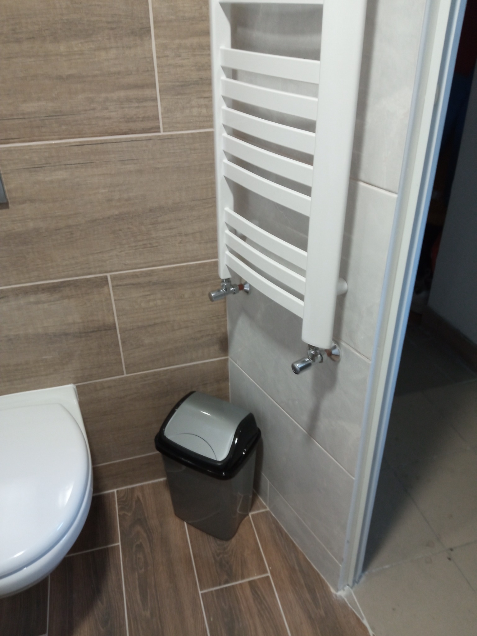 Fragment łazienki z grzejnikiem drabinkowym, płytkami imitującymi drewno na ścianie i podłodze, oraz koszem na śmieci z szarą pokrywą.