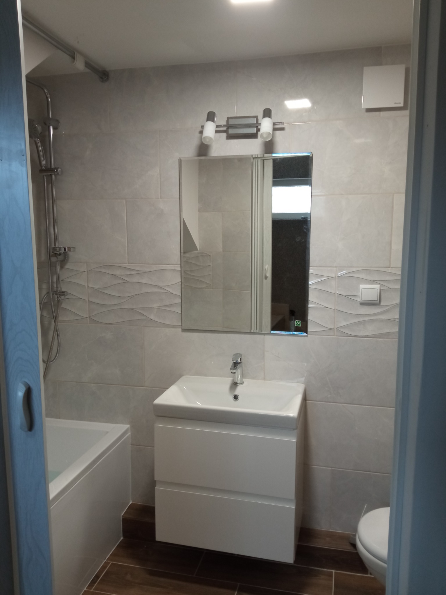 Wykończona łazienka w odcieniach szarości z brązowymi płytkami podłogowymi, obejmująca wannę z prysznicem, umywalkę z szafką, lustro z oświetleniem i toaletę.