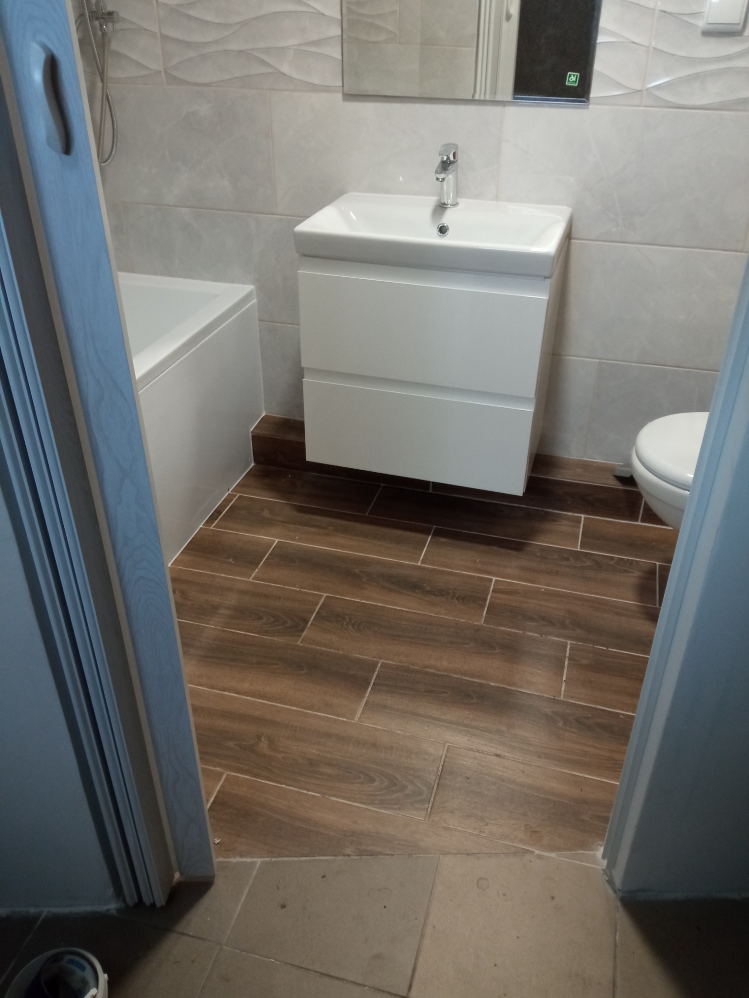 Wykończona łazienka z brązowymi płytkami imitującymi drewno, białą umywalką z szafką, lustrem i częściowo widoczną wanną, sfotografowana zza uchylonych drzwi.