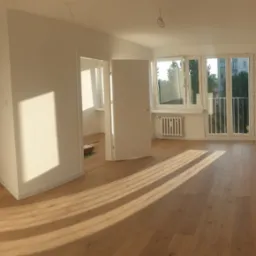 Jasne, puste wnętrze mieszkania po remoncie z drewnianą podłogą, białymi ścianami, oknami balkonowymi i otwartymi drzwiami do drugiego pomieszczenia.