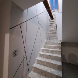 Stanislav Timonov - Klatka schodowa w trakcie wykończenia: ściany z dekoracyjną szpachlą w geometryczne wzory, surowe betonowe schody, drewniana drabina oparta o ścianę, widoczne puszki elektryczne.