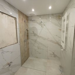 Stanislav Timonov - Wnętrze łazienki w trakcie prac wykończeniowych: białe płytki imitujące marmur, fragment ściany przygotowany pod zabudowę, zamontowana kabina prysznicowa z deszczownicą i grzejnik drabinkowy.