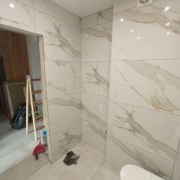 Stanislav Timonov - Łazienka w trakcie wykańczania z białymi płytkami imitującymi marmur, widoczna drabina, wiadro i częściowo zamontowana toaleta.