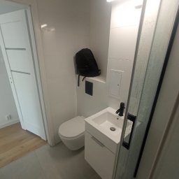 Stanislav Timonov - Nowoczesna, minimalistyczna łazienka z białymi płytkami, podwieszaną toaletą, szafką z umywalką i czarną armaturą; plecak leżący na półce nad toaletą.