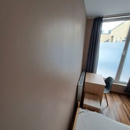 Stanislav Timonov - Ściana w pokoju pomalowana na ciepły, beżowy kolor, widoczny grzejnik, biurko z krzesłem przy oknie z zasłoną i częściowo widoczne łóżko.