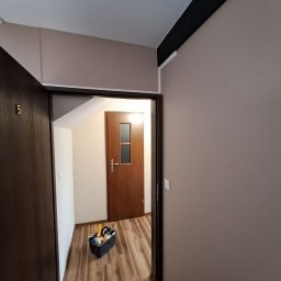 Stanislav Timonov - Korytarz po odświeżeniu: świeżo pomalowane ściany w odcieniach beżu, widoczna skrzynka z narzędziami malarskim przy wejściu do mieszkania z brązowymi drzwiami.