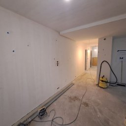 Stanislav Timonov - Wnętrze w trakcie prac wykończeniowych: świeżo wyszpachlowane ściany z zaznaczonymi punktami, widoczne przewody elektryczne, rury przy podłodze, odkurzacz przemysłowy i plany architektoniczne...