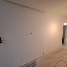 Stanislav Timonov - Wnętrze w trakcie prac wykończeniowych: wygładzone ściany z widocznymi otworami na gniazdka elektryczne zabezpieczone taśmą malarską, surowa podłoga, widok na kolejne pomieszczenia przez otwory...