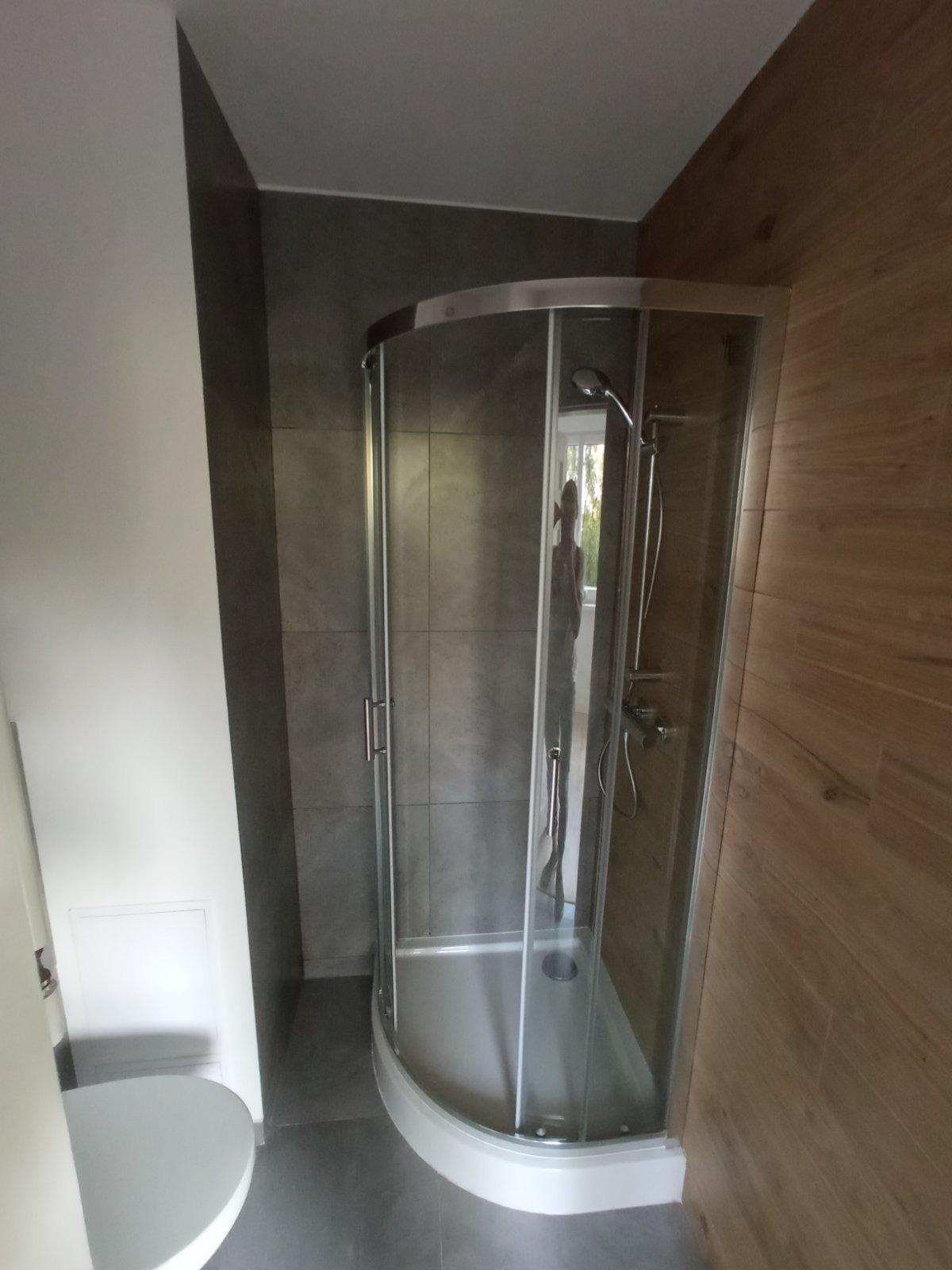 Narożna kabina prysznicowa z chromowanymi profilami i brodzikiem, umieszczona w łazience z szarymi płytkami ściennymi i panelami imitującymi drewno, widoczne częściowo białe elementy armatury.