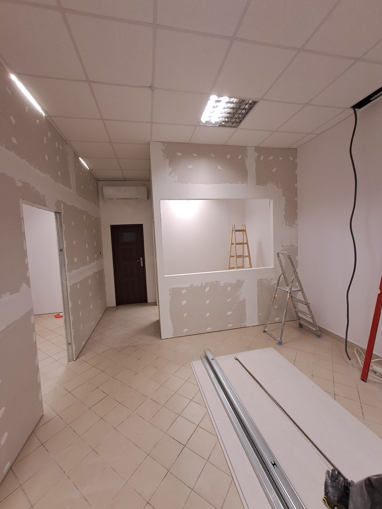 Wnętrze w trakcie prac przygotowawczych do wykończenia: ściany z płyt kartonowo-gipsowych po szpachlowaniu, widoczne profile metalowe i drabina w otworze okiennym, sufit kasetonowy.