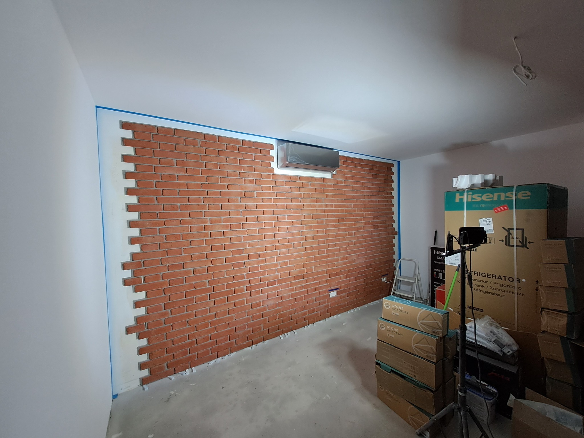 Ściana wyłożona cegłą dekoracyjną w trakcie montażu, z widocznymi elementami mocującymi, zabezpieczona niebieską taśmą malarską, z zamontowanym klimatyzatorem, w pomieszczeniu z kartonami i sprzętem.