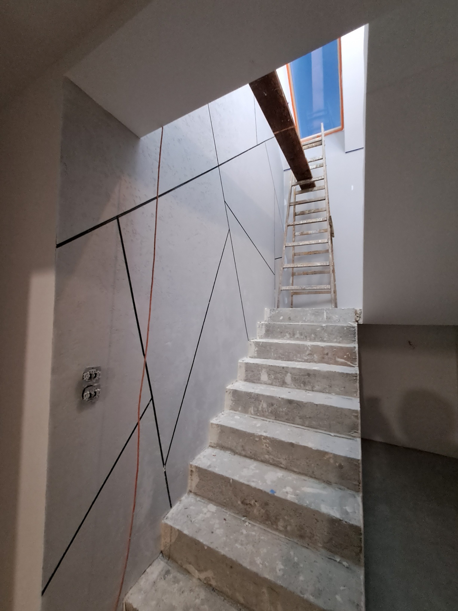 Klatka schodowa w trakcie wykończenia: ściany z dekoracyjną szpachlą w geometryczne wzory, surowe betonowe schody, drewniana drabina oparta o ścianę, widoczne puszki elektryczne.