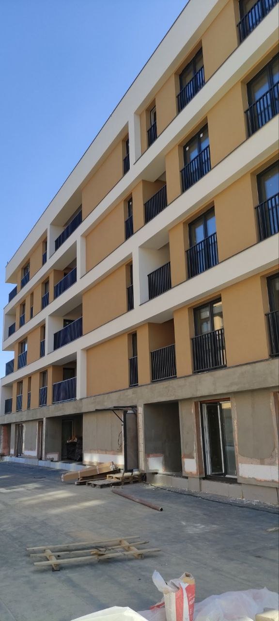 Wielopiętrowy budynek mieszkalny w trakcie wykończenia elewacji, widoczne balkony z czarnymi barierkami, częściowo otynkowane ściany na parterze, palety i materiały budowlane na zewnątrz.