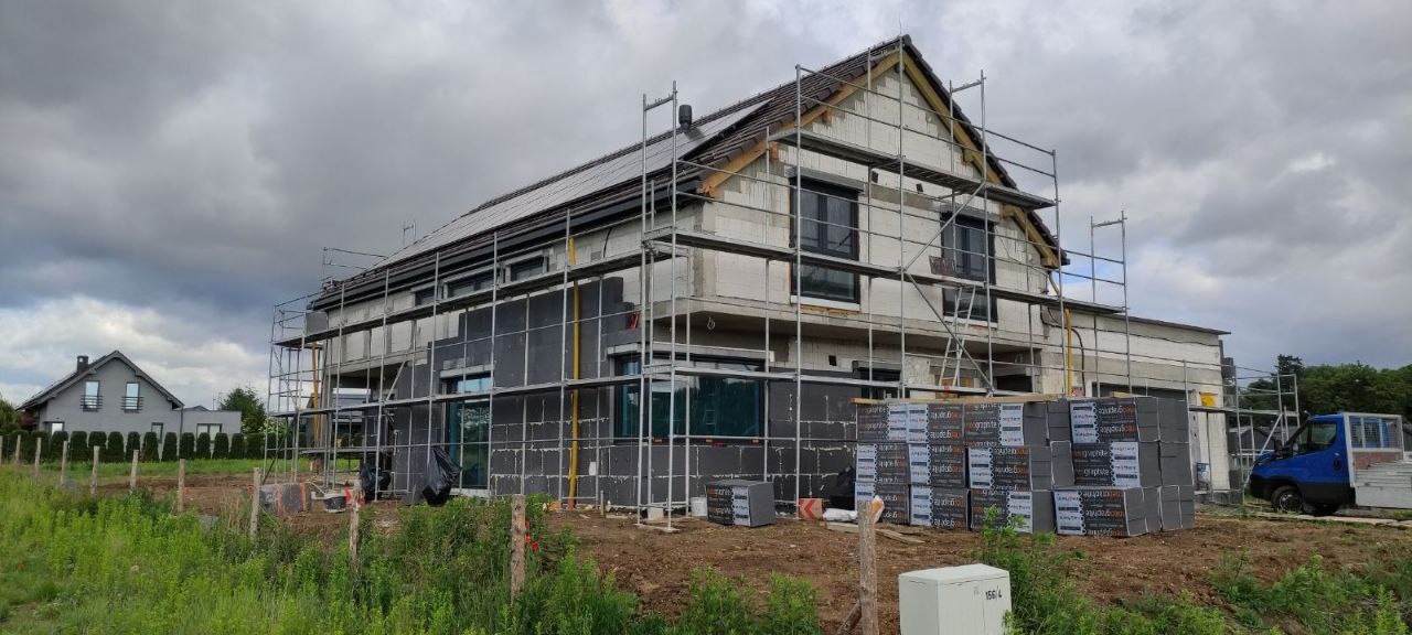 Dom w trakcie ocieplania elewacji, widoczne rusztowania, częściowo nałożony styropian grafitowy, palety z materiałem izolacyjnym, pochmurne niebo.