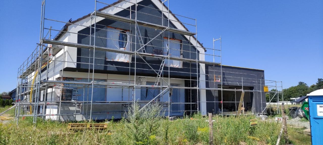 Dom jednorodzinny w trakcie ocieplania elewacji zewnętrznej, widoczne rusztowania i zabezpieczone okna folią, tynk strukturalny w kolorze antracytowym na części ściany, trawa i chwasty na pierwszym...