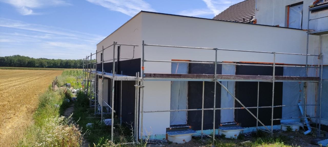 Dom jednorodzinny w trakcie ocieplania elewacji, widoczne rusztowania, czarne płyty izolacyjne i częściowo otynkowana ściana w kolorze białym na tle pola i lasu.