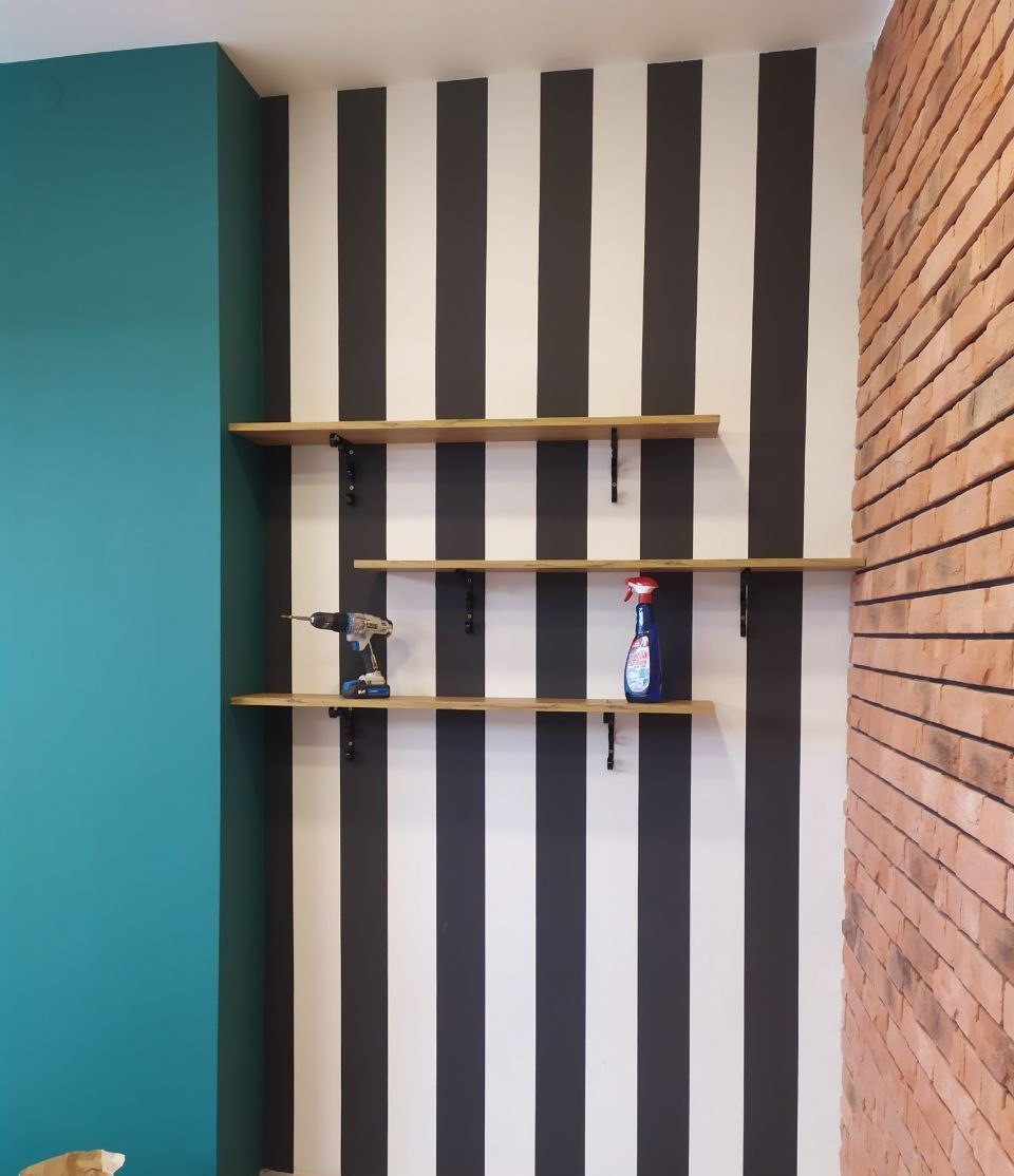 Ściana w biało-czarne pasy z trzema drewnianymi półkami na czarnych wspornikach, na których leżą wiertarka akumulatorowa i butelka płynu do czyszczenia, obok ściana z cegły i turkusowa ściana.