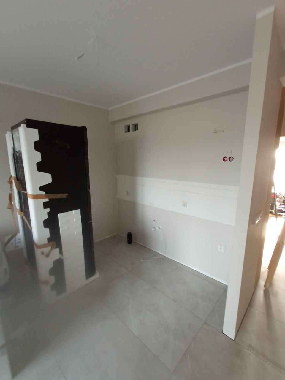 Wnętrze w trakcie wykańczania: szare płytki podłogowe, fragment ściany z elementami instalacji elektrycznej, mebel zabezpieczony folią i styropianem, widoczne niedociągnięcia na suficie.