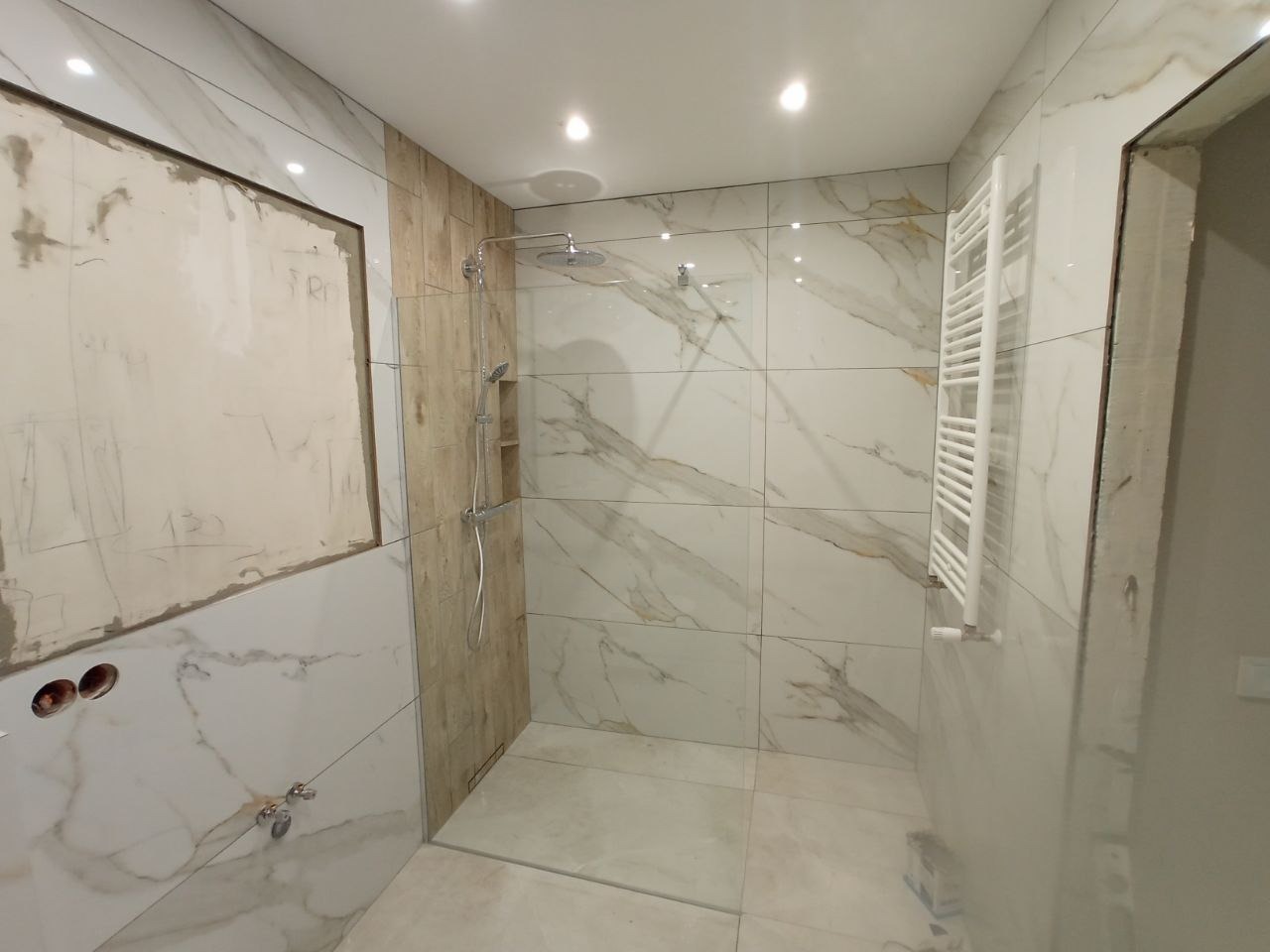 Wnętrze łazienki w trakcie prac wykończeniowych: białe płytki imitujące marmur, fragment ściany przygotowany pod zabudowę, zamontowana kabina prysznicowa z deszczownicą i grzejnik drabinkowy.