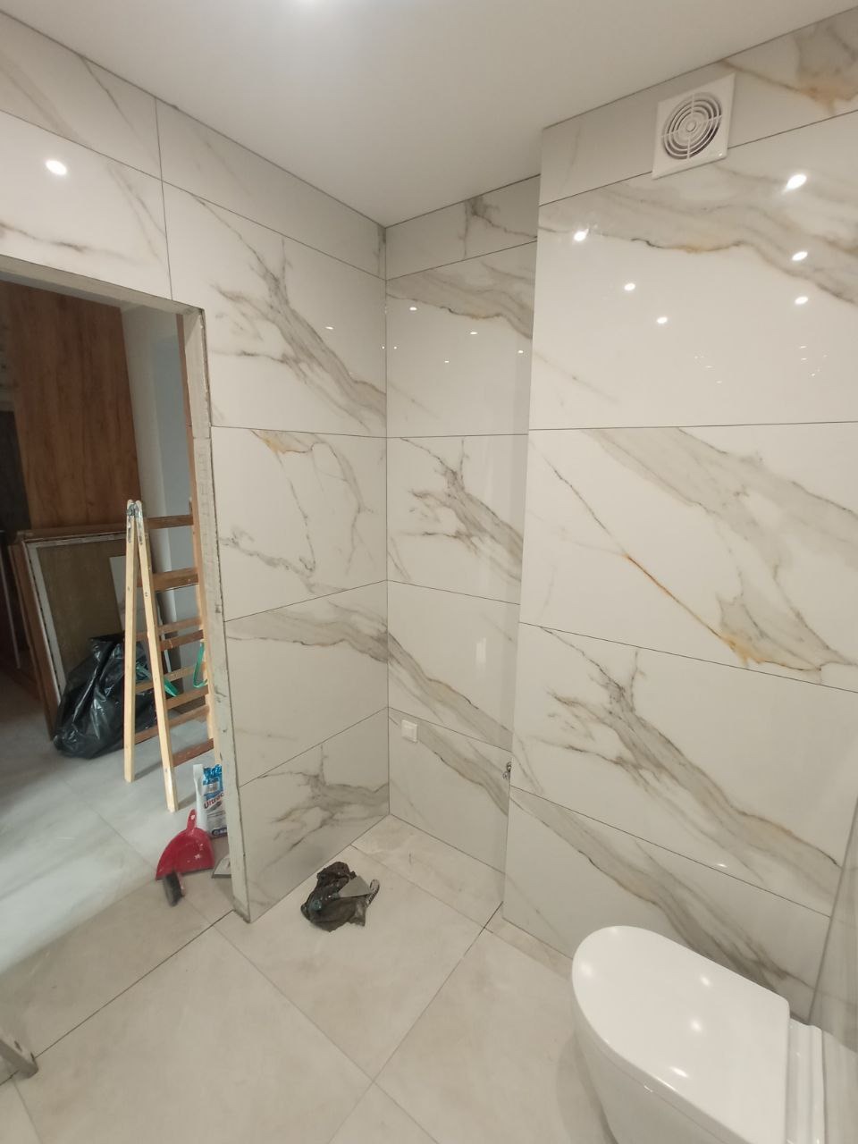 Łazienka w trakcie wykańczania z białymi płytkami imitującymi marmur, widoczna drabina, wiadro i częściowo zamontowana toaleta.